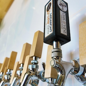 以下が含まれることがあります： 木製のハンドルが付いたビールタップの列のクローズアップ。前面のタップには、「Midnight Drip Coffee」と「Oak & Vine Collective」という文字が印刷された黒い長方形のトップが付いています。タップは光沢のある金属製です。