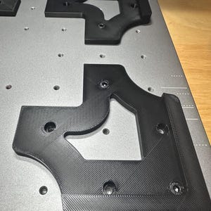 May include: Black, geometric-shaped components with screw holes are arranged on a gray surface. The components have a textured, ridged pattern. The arrangement suggests a mechanical or industrial application.