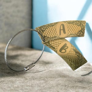 May include: Gold-colored luggage tags with the letters "A" and "B" printed on them. The tags have a fish scale pattern and decorative scrollwork. They are attached to a silver-colored wire loop.
