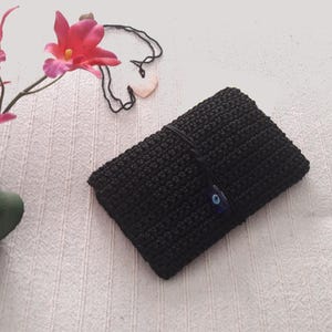 Könnte beinhalten: Schwarze gehäkelte Tasche mit einem blauen bösen Blick-Anhänger. Die Tasche ist aus feinem, strukturiertem Garn gefertigt und hat ein schlichtes, elegantes Design.