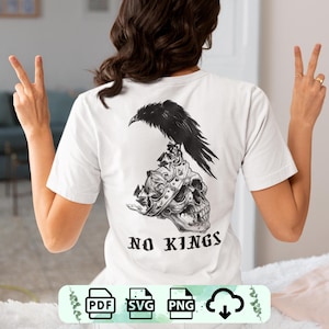 May include: White t-shirt featuring a black raven perched atop a skull wearing a crown. The phrase "NO KINGS" is printed below the skull. The design is centered on the back of the shirt.