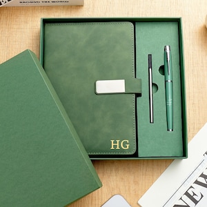 Puede incluir: Juego de diario y bolígrafo de cuero verde en una caja de regalo. El diario tiene un cierre plateado y las iniciales "HG" en relieve. Se incluye un bolígrafo a juego y un bolígrafo delgado. El juego se presenta sobre una superficie de madera.