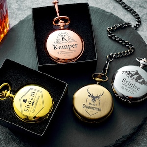 May include: Four engraved pocket watches in gold, silver, and copper tones, each with a chain. The watches are displayed on a dark gray surface. One watch is in a black box. The watch faces feature custom text and designs.