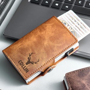 Peut inclure: Un portefeuille en cuir marron avec un logo de tête de cerf et le nom "COLLIN" gravé sur le devant. Le portefeuille est ouvert, révélant plusieurs billets. Le portefeuille est à côté d'un ordinateur portable.