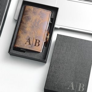 Puede incluir: Una cartera de cuero marrón con las iniciales "AB" grabadas en la parte delantera, presentada en una caja de regalo negra. También se ven un bolígrafo plateado y un cuaderno negro, junto a un ordenador portátil. La cartera tiene un aspecto de cuero envejecido.