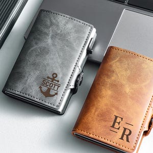 May include: Two leather cardholders are shown, one grey and one brown. The grey cardholder has an anchor design with the name "SCOTT" below. The brown cardholder features the initials "E R". Both have a metal frame and a push-button mechanism.