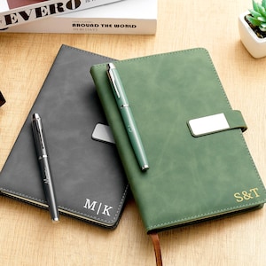 Pode incluir: Dois cadernos personalizados, um cinza e outro verde, com canetas a condizer. O caderno cinza tem as iniciais M|K, e o caderno verde tem S&T. Ambos os cadernos têm uma caneta em cima e uma fita de marcador.