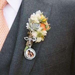 以下が含まれることがあります： グレーのスーツジャケットに取り付けられた、カップルの写真が入った小さな丸い銀色のロケットが付いたブートニア。ブートニアは、白、黄色、オレンジの花と緑と灰色の多肉植物のクラスターが特徴です。 桃色のネクタイが見えます。