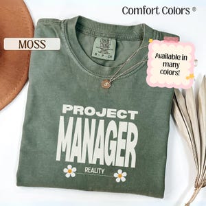 May include: A moss green t-shirt with the words "PROJECT MANAGER REALITY" in white. The shirt has a crew neck and short sleeves. A silver necklace with a pendant is on the shirt. The shirt is from Comfort Colors.