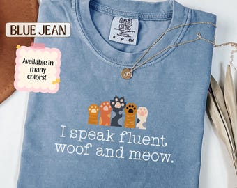 May include: A blue jean colored t-shirt with a graphic of five colorful animal paws and the white text I speak fluent woof and meow. The shirt features a Comfort Colors brand label at the neckline and is paired with a gold chain necklace.