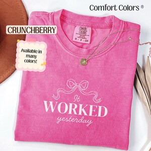 May include: A folded, bright pink Comfort Colors t-shirt with the text "It WORKED yesterday" in white. A gold necklace with a pendant rests on the shirt. The shirt is labeled "Crunchberry" and is available in many colors.