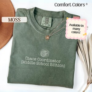 May include: A moss green Comfort Colors t-shirt with the text "Chaos Coordinator (Middle School Edition)". The shirt has a crew neck and short sleeves. A silver necklace with a pendant is on the shirt. The text "Available in many colors!" is also visible.