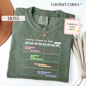 May include: A moss green crew neck t-shirt featuring a white graphic of a Gantt chart with the text Keeping Everyone on Track. The shirt has a Comfort Colors brand label at the neck and is displayed with a gold chain necklace resting on the fabric.
