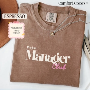 May include: Espresso-colored t-shirt with the words "Project Manager Club" in white and pink. The shirt is a Comfort Colors brand, and the tag indicates it is a size small. A silver necklace with a pendant is draped across the shirt.