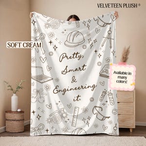 May include: A soft cream-colored plush blanket featuring a dark brown line-art pattern of engineering-themed icons including a hard hat, laptop, gears, compass, ruler, and safety goggles, with the text "Pretty, Smart & Engineering it."