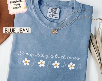 May include: A blue jean Comfort Colors t-shirt with the text "It's a good day to teach music" in white. The shirt features musical notes and daisy graphics. A gold necklace with a pendant rests on the shirt.