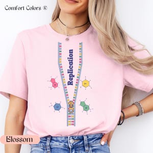 May include: Pink t-shirt with a graphic of a zipper representing DNA replication. The zipper is open and the DNA strands are visible. The graphic includes the letters A, T, C, and G, which represent the four nitrogenous bases in DNA. The text "Replication" is written vertically on the zipper.