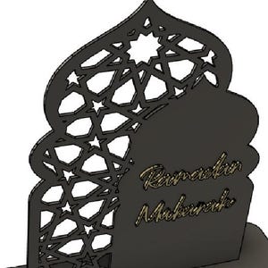 May include: A black laser-cut Ramadan Mubarak decoration. The design features an arched silhouette with geometric star and polygon cutouts. The words "Ramadan Mubarak" are in a gold script font.