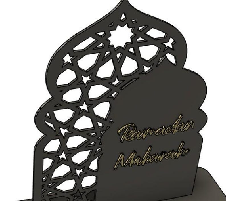 May include: A black laser-cut Ramadan Mubarak decoration. The design features an arched silhouette with geometric star and polygon cutouts. The words "Ramadan Mubarak" are in a gold script font.