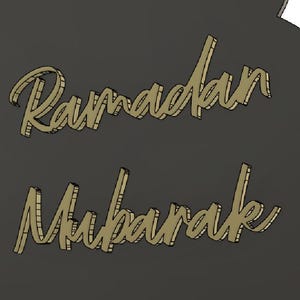 May include: A dark grey decorative piece with the words "Ramadan Mubarak" in gold script. The design includes a geometric star pattern on the left side. The text is in a stylized, elegant font.