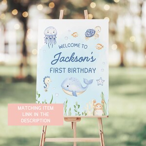 May include: Welcome to Jackson's First Birthday sign featuring a blue whale, a seahorse, a jellyfish, a turtle, and a crab on a blue background with white bubbles.