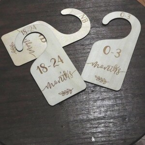 May include: Wooden baby clothes dividers with engraved text. The dividers are light brown and feature the age ranges "Newborn", "0-3 months", and "18-24 months". Each divider has a curved top for hanging and decorative leaf accents.