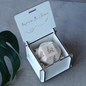 May include: A white wooden box with an open lid, engraved with "Aaron & Jenn" and a heart and airplane symbol. Inside, a wooden dodecahedron with engraved figures rests on shredded paper.