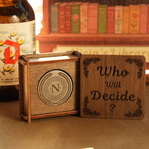 May include: Wooden box with a silver emblem featuring the letter "N" inside a laurel wreath. A wooden coaster with the words "Who will Decide?" is next to the box. The box and coaster are on a brown surface.