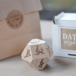 May include: A wooden, multi-sided die with engraved illustrations of various activities. The die is light brown and sits on a light surface. A white box with the words "DATE night" is in the background, along with a brown paper bag.