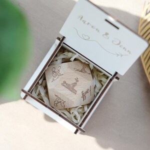 May include: A wooden dice with various positions engraved on each side, presented in a white wooden box. The box lid is engraved with the names "Aaron & Denn" and a heart symbol. The dice is nestled in shredded paper.