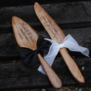 May include: Wooden cake server and knife set with engraved names and date. The server has a black bow, and the knife has a white bow. The names "Meriah & Daniel" and the date "05.06.26" are engraved on the server.