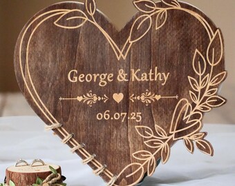 Könnte beinhalten: Ein herzförmiges Hochzeitszeichen aus Holz mit den eingravierten Namen "George & Kathy" und dem Datum "06.07.25". Das Schild hat ein Blätterdesign und eine Schnur-Umrandung. Ein kleiner Holzringhalter mit zwei Ringen befindet sich im Vordergrund.