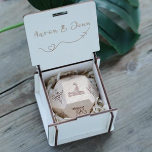 May include: A wooden box with the names "Aaron & Jenn" engraved on the lid, along with a heart and airplane symbol. Inside, a wooden dodecahedron with various illustrations rests on wood shavings. The box is open, revealing the contents.