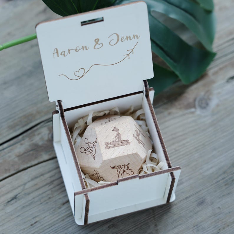 May include: A wooden box with the names "Aaron & Jenn" engraved on the lid, along with a heart and airplane symbol. Inside, a wooden dodecahedron with various illustrations rests on wood shavings. The box is open, revealing the contents.