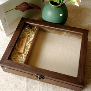 Könnte beinhalten: Eine rechteckige dunkelbraune Holz-Präsentationsbox mit einem klaren Glasdeckel und einem Metallverschluss. Das Innere verfügt über einen beigen Stoffboden und ein separates Fach mit hellen Holzspänen, das einen kleinen Holz-USB-Stick mit der Gravur Daniel enthält.