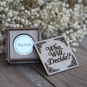 May include: A square wooden box with a sliding lid featuring the engraved text Who Will Decide? and floral corner accents. Inside, a circular silver-toned metal decision coin is visible, inscribed with the name Sarah and the text Decision Coin Flip to Decide.