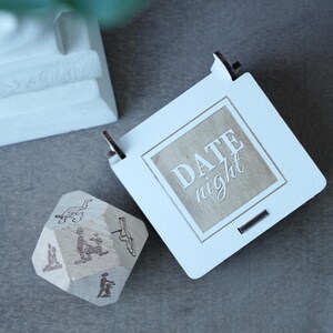 May include: A white wooden box with the words "DATE night" engraved on it, next to a wooden die with various illustrations. The box has a square shape with a light brown interior. The die has different illustrations on each side.