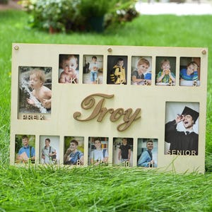 以下が含まれることがあります： 複数の小さな写真の開口部と筆記体の「Troy」という名前が付いた木製のフォトフレーム。フレームには、幼稚園から最終学年まで、さまざまな年齢の子供の写真が収められています。フレームは緑の草の表面にあります。