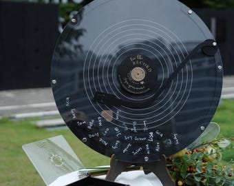 Könnte beinhalten: Ein kreisförmiges Gästebuch aus transparentem Acryl in Form einer Schallplatte mit einem schwarzen Mittellabel, das den Text "For the Record" und "sign your name" trägt. Es enthält einen schwarzen Tonarm, einen Holzständer und eine Box mit schwarzen Marken.
