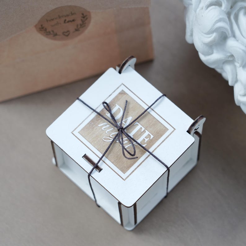 May include: A white wooden box tied with dark twine. The box has a square shape with a lid that features the words "DATE night" on a light brown background. A brown paper bag with a "handmade with love" sticker is in the background.