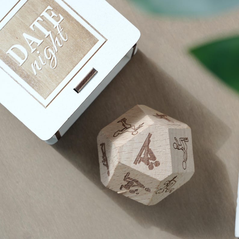 May include: A wooden date night dice with engraved activity icons and a white box with the words "DATE night" on the lid. The dice is light brown and the box is white.