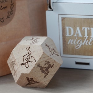 May include: A wooden decahedron die with etched illustrations of various sexual positions. The die is light brown and sits in front of a white box with the words "DATE night" on it.