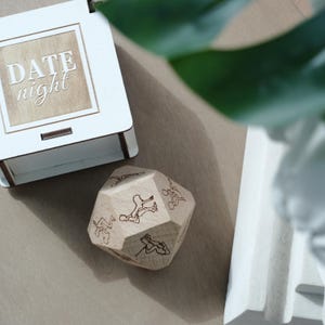 May include: A wooden date night box with the words "DATE night" engraved on the lid. A multi-sided wooden die with engraved illustrations of couples is next to the box. The box and die are on a light brown surface.