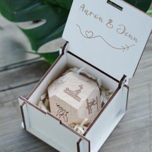 May include: A wooden box with the names "Aaron & Jenn" engraved on the lid. Inside, a wooden geometric shape with various illustrations rests on shredded paper. The box is open, revealing the contents.