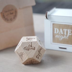 May include: A wooden, multi-sided die with etched illustrations of various activities. The die is light-colored and sits in front of a white box with the words "DATE night" printed on it. A brown paper bag is in the background.