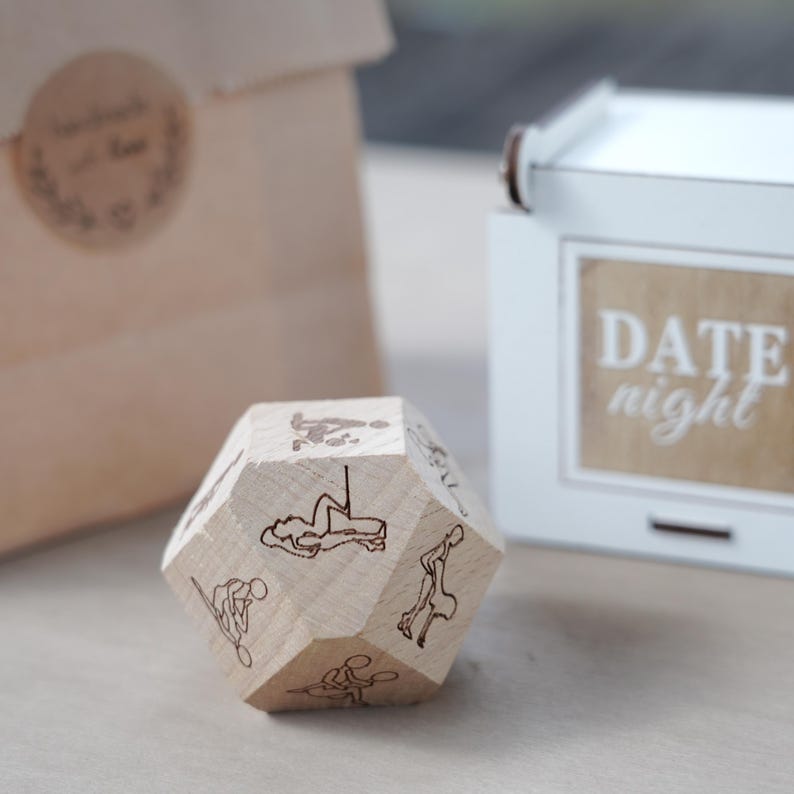 May include: A wooden, multi-sided die with etched illustrations of various activities. The die is light-colored and sits in front of a white box with the words "DATE night" printed on it. A brown paper bag is in the background.