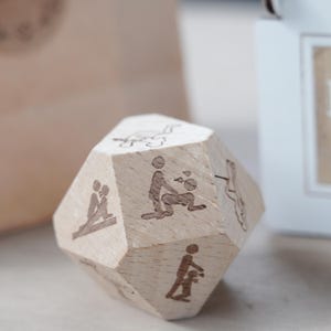 May include: A wooden, multi-sided die with etched illustrations depicting various sexual positions. The die is made of light-colored wood and has a natural finish. The illustrations are simple line drawings.