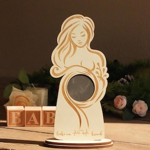 May include: Wooden photo frame in the shape of a pregnant woman. The frame has a circular opening for a photo and is engraved with the phrase "liebe im bauch" and a heartbeat symbol. The frame is made of light-colored wood.