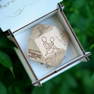 Peut inclure: Un dé en bois à plusieurs faces avec des illustrations gravées de couples dans diverses positions intimes, présenté à l'intérieur d'une boîte blanche ouverte. La boîte a un cœur décoratif et le mot "Marry" gravé sur le couvercle.