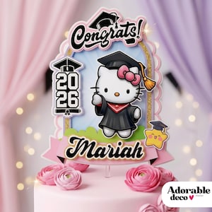 Puede incluir: Adorno para tarta rosa con Hello Kitty vestida con toga de graduación, birrete y lazo rosa. El adorno dice "Congrats!" y "Mariah" con el año "2026". La tarta está decorada con rosas rosas.